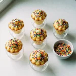 Five viral dot cakes in clear cocktail glasses covered in rainbow nonpareil sprinkles on white marble