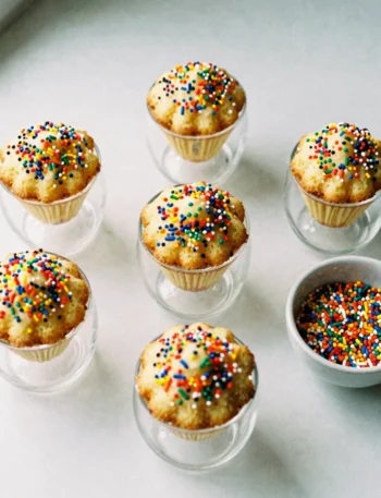 Five viral dot cakes in clear cocktail glasses covered in rainbow nonpareil sprinkles on white marble